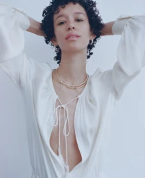 Model with dark curly hair wearing a white silk blouse and gold jewelry against a seamless background for Present Space.