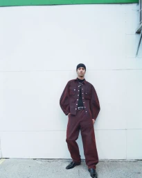 Man wearing a burgundy button-down shirt and matching trousers with a black beanie against a white wall.