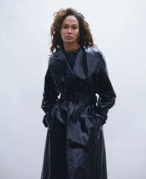 Joan Smalls wearing a glossy black belted trench coat against a plain white background