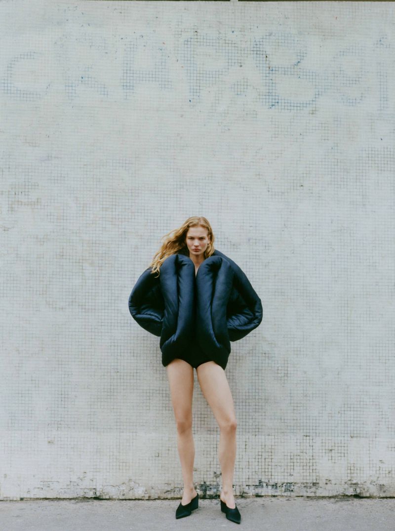 Full-length shot of a woman in a dark puffer jacket and black mules posing against a textured tile wall in an outdoor setting.