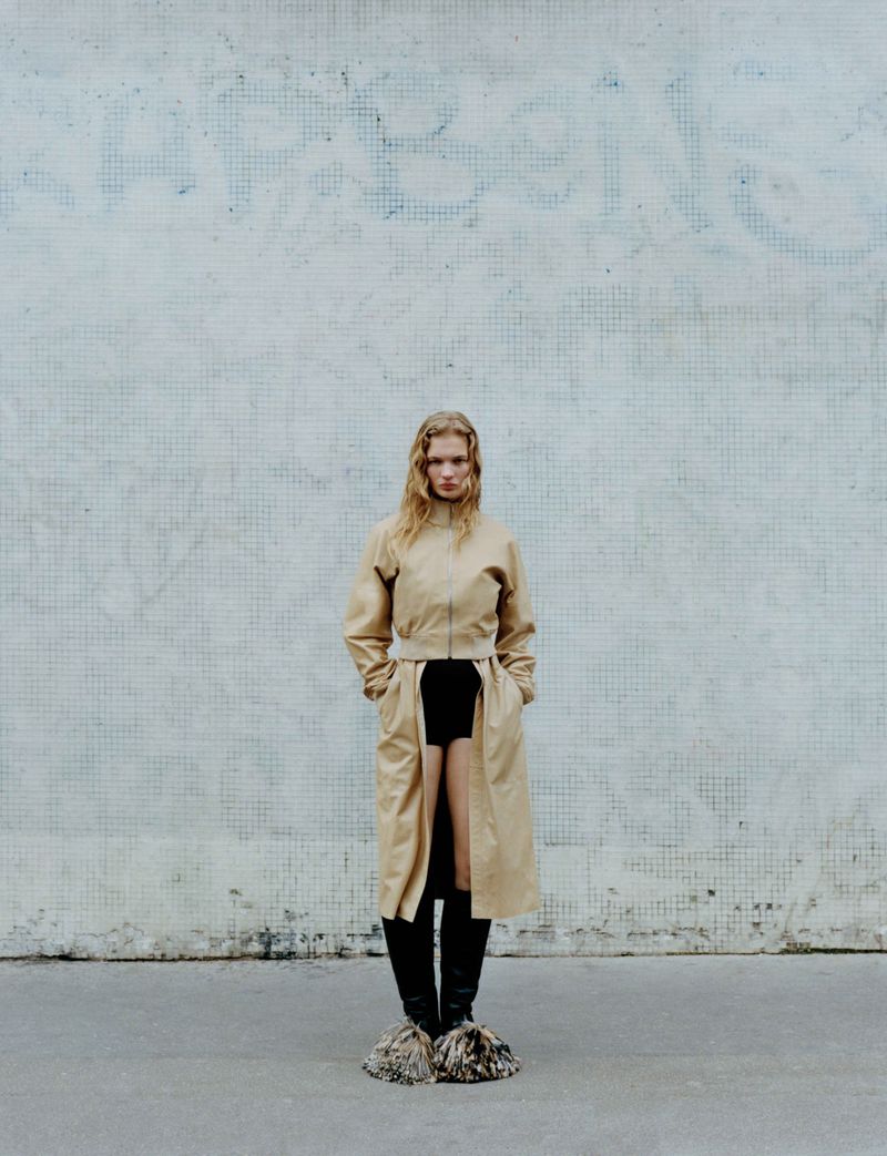 A full-length shot of a blonde adult female fashion model in a long beige coat and black boots, standing against a tiled wall.
