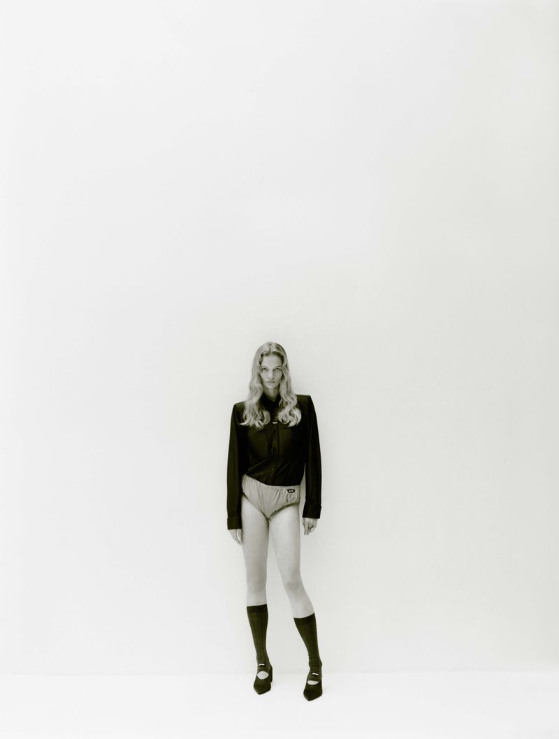 Model standing against a plain white background wearing a dark jacket, light shorts, knee socks, and flat shoes.