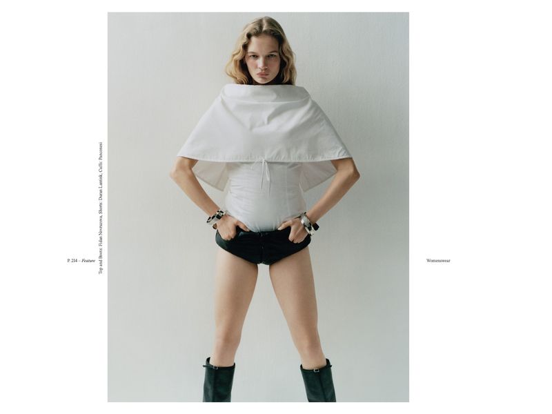 Fashion editorial shot of an adult female model with blond hair wearing a white shirt, black shorts, and black boots against a white background.