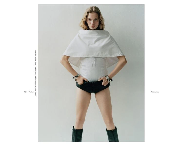 Fashion editorial shot of an adult female model with blond hair wearing a white shirt, black shorts, and black boots against a white background.
