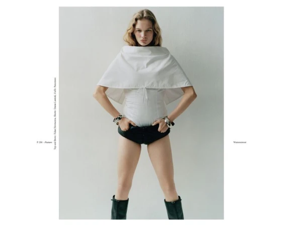 Fashion editorial shot of an adult female model with blond hair wearing a white shirt, black shorts, and black boots against a white background.