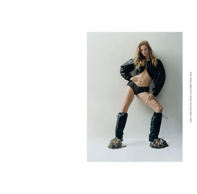 Fashion editorial shot of a blonde woman in a black leather jacket, black shorts, and tall black boots with fur accents, against a white wall.