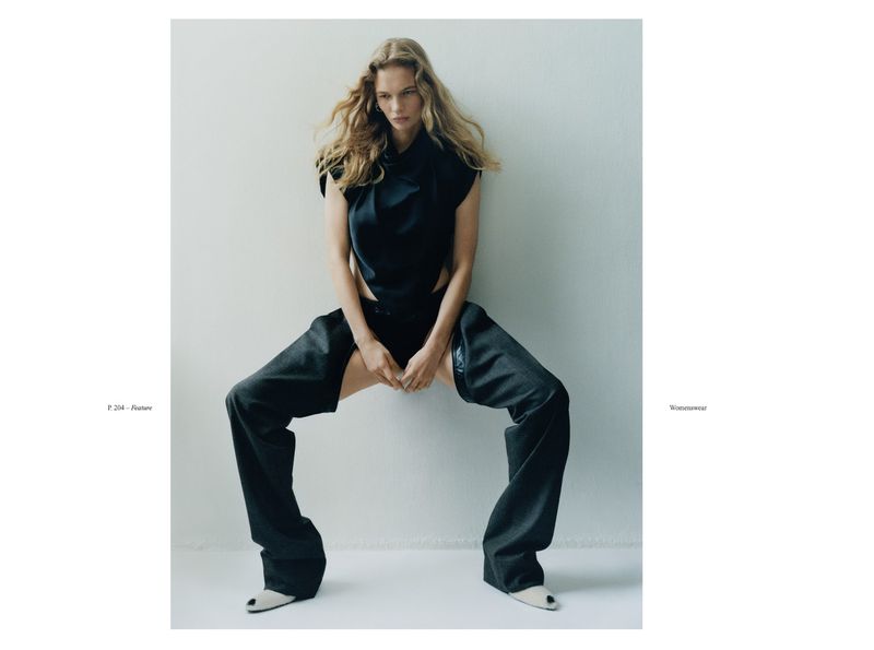 A woman in a black top and open-legged pants poses against a white wall in a fashion studio, showcasing a modern, edgy look.