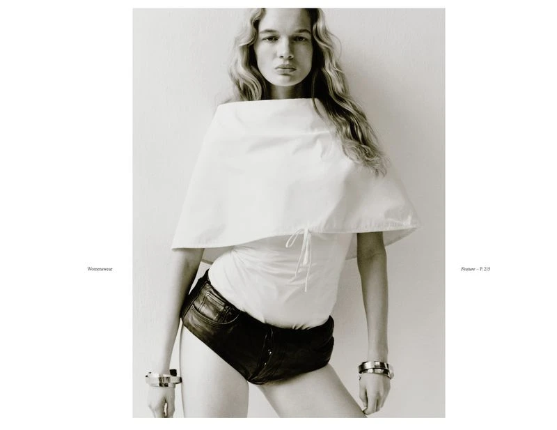 Portrait of a female model in a white cape top and black shorts, wearing silver bracelets, posing against a neutral background in a studio.