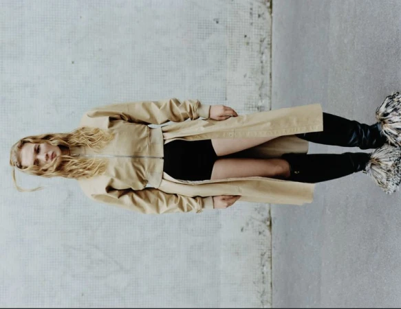 Model wearing a tan coat and black boots against a concrete wall for Present Space