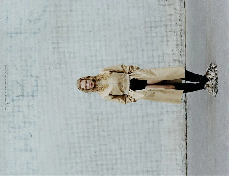 Fashion shot of a woman in a tan coat standing on a concrete floor in front of a textured wall.  Editorial style.
