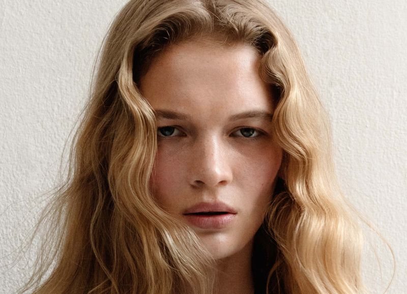 Close-up portrait of a blonde woman with fair skin and light eyes, against a neutral background, showcasing beauty and fashion.