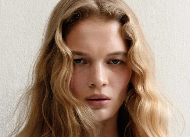 Close-up portrait of a blonde woman with fair skin and light eyes, against a neutral background, showcasing beauty and fashion.