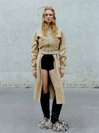 A model stands outdoors against a textured wall, wearing a tan jacket and trench coat, black shorts, and distinctive furry boots in a fashion shot.
