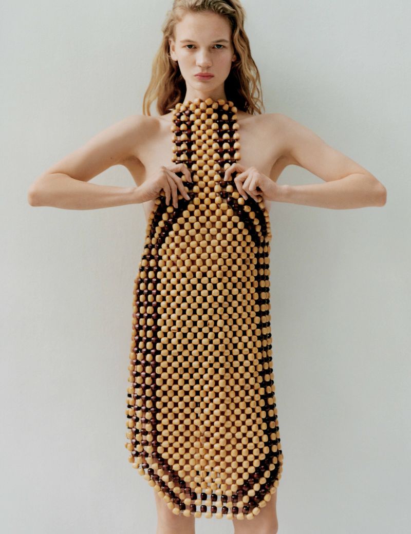 Young fair-skinned model in a wood-beaded dress against a white wall, fashion-forward and eye-catching. Studio shot with natural light.