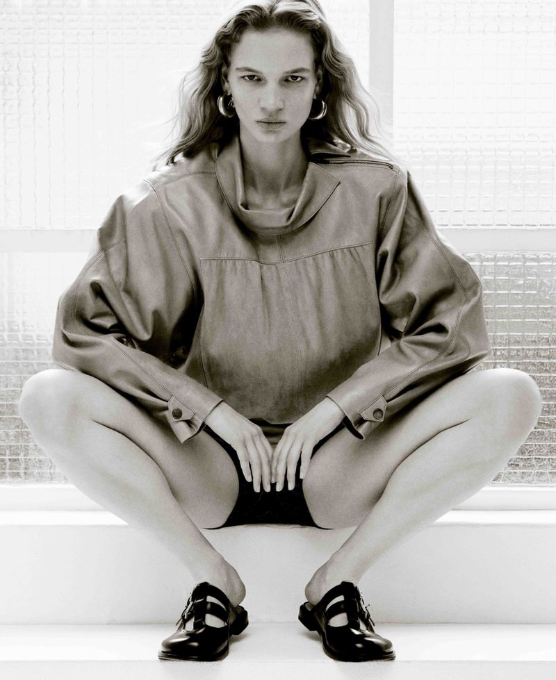 Monochrome shot of a woman in a tan top and black shorts, seated with legs apart and hands between her legs, against a white background.