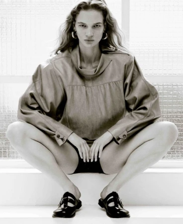 Monochrome shot of a woman in a tan top and black shorts, seated with legs apart and hands between her legs, against a white background.