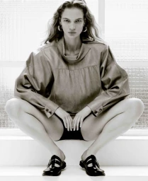 Model squatting in a cowl neck top and black shoes against a grid window background