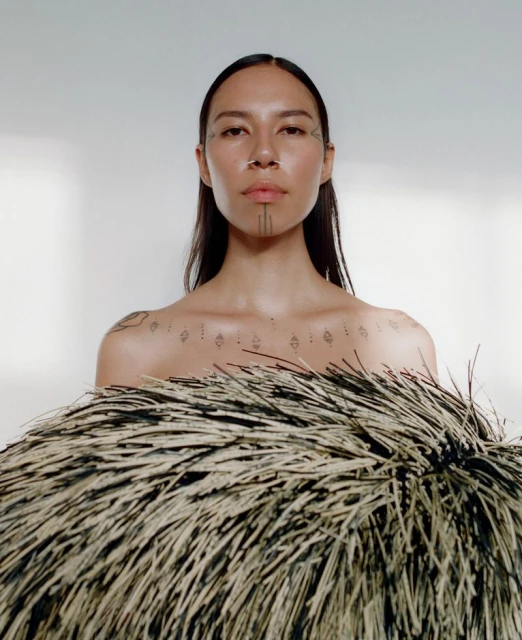 Portrait of a woman with face and body tattoos against a white background, featuring a fringed textile obscuring part of her torso.