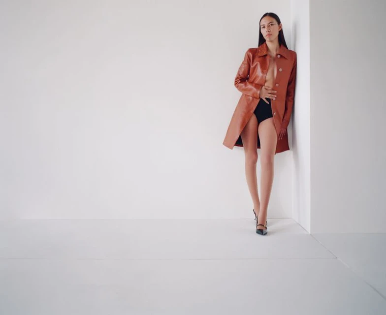 Adult woman in stylish orange coat and black heels poses against a white wall in a studio, fashion-forward and confident.