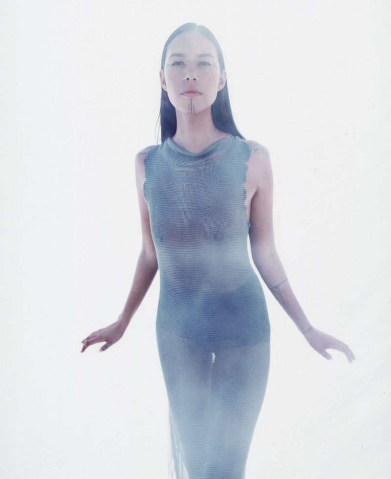 Woman in sheer blue dress poses with arms slightly outstretched against a bright white background in a fashion photography style.