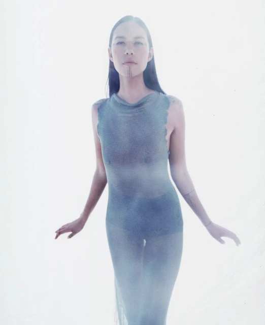 Woman in sheer blue dress poses with arms slightly outstretched against a bright white background in a fashion photography style.
