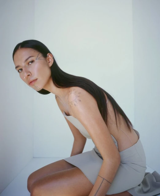 An adult female with long dark hair and face tattoos poses in a beige dress against a white studio backdrop in a medium indoor shot.
