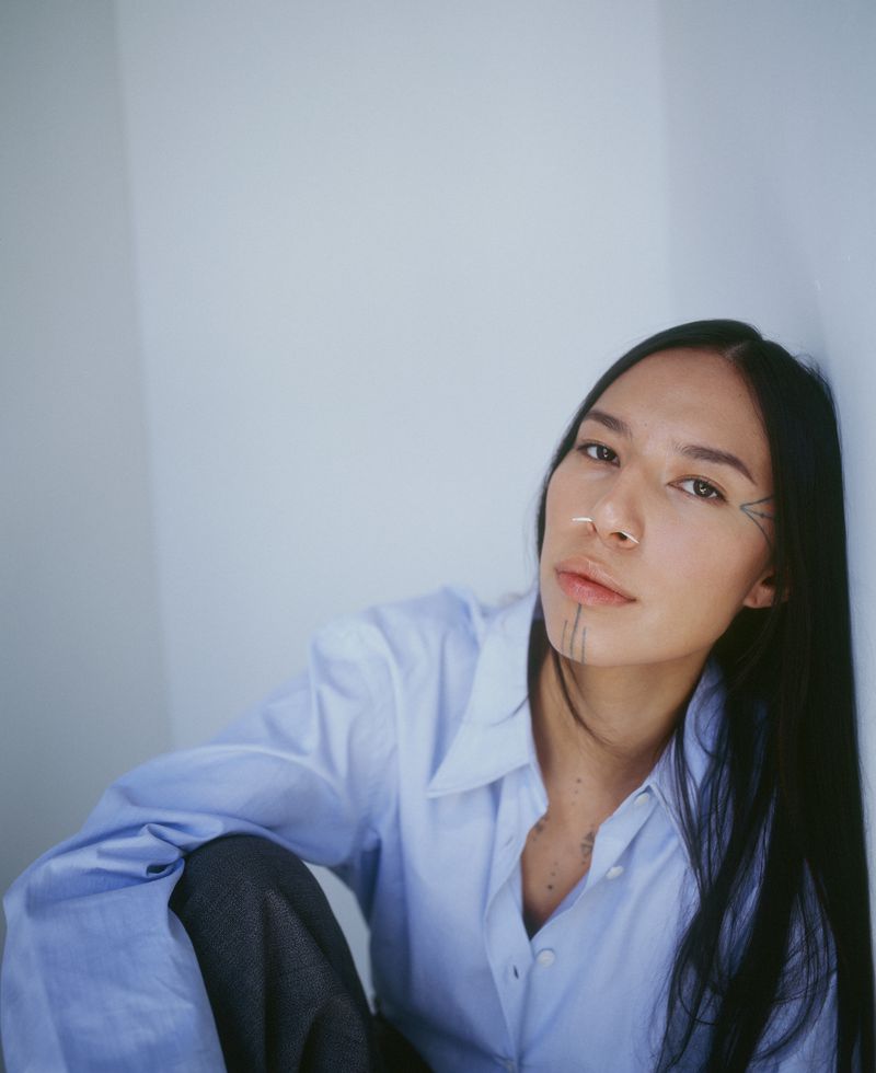 A young adult woman with face tattoos poses leaning against a wall. She is wearing a blue button-down shirt and grey pants.