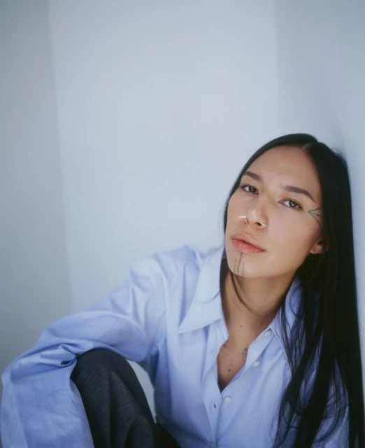 A young adult woman with face tattoos poses leaning against a wall. She is wearing a blue button-down shirt and grey pants.