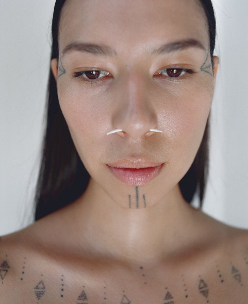 Close-up portrait of an adult female with tribal-style tattoos on her face, chin, and chest, wearing labret piercings, against a white backdrop.