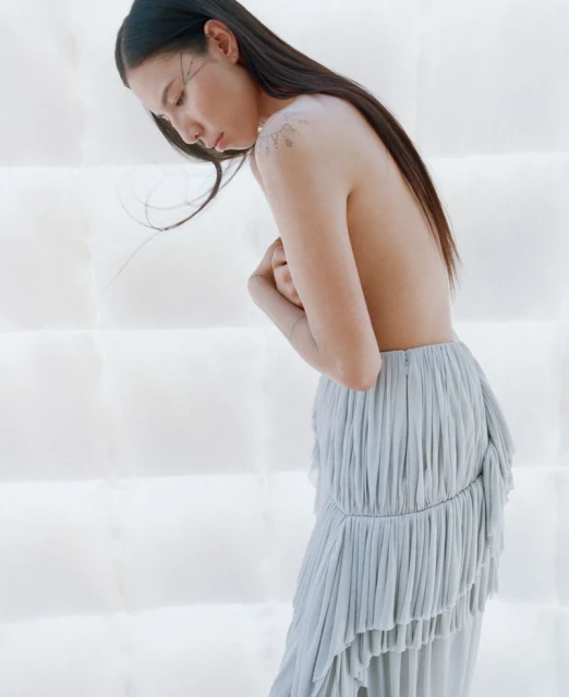 Profile of a woman with long dark hair in a flowing gray skirt, covering her chest with her arms in front of a white background.