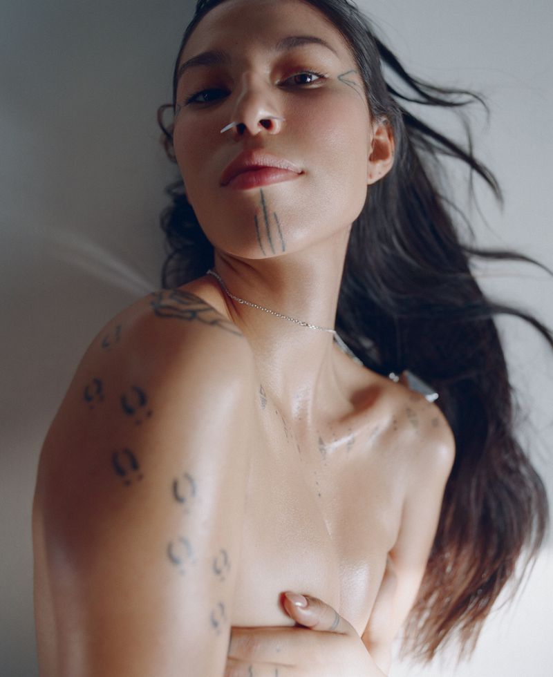 Close-up portrait of a woman with traditional facial tattoos, long dark hair, and minimal jewelry, set against a soft, diffused background.