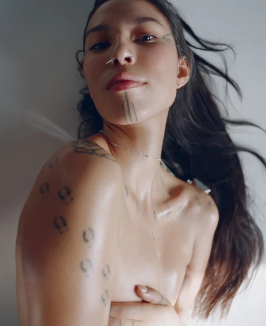 Close-up portrait of a woman with traditional facial tattoos, long dark hair, and minimal jewelry, set against a soft, diffused background.