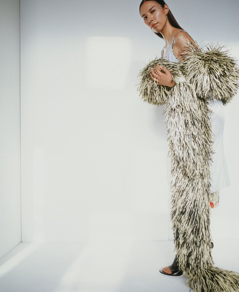 A young woman in a white dress poses in a photo studio, wearing a long, tan and black shaggy coat that extends to the floor.