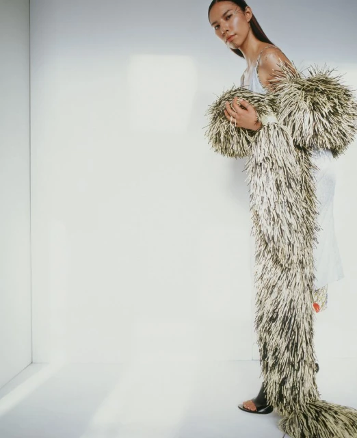 A young woman in a white dress poses in a photo studio, wearing a long, tan and black shaggy coat that extends to the floor.