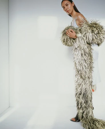 A young woman in a white dress poses in a photo studio, wearing a long, tan and black shaggy coat that extends to the floor.