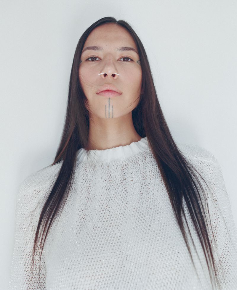 Portrait of an Indigenous woman with long dark hair wearing a textured white sweater, posing against a white studio background.