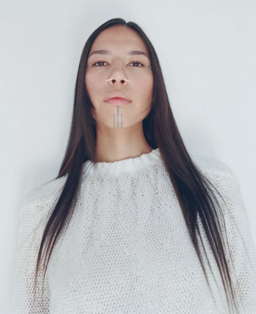Portrait of an Indigenous woman with long dark hair wearing a textured white sweater, posing against a white studio background.