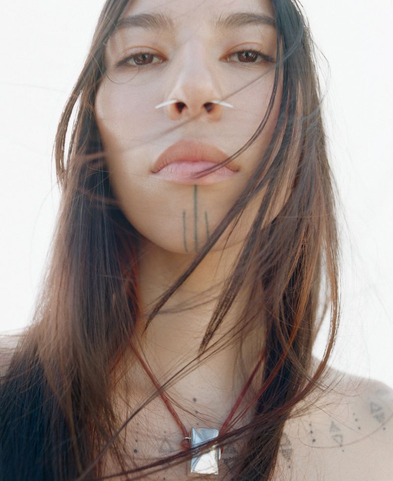 Close-up portrait of a young adult woman with long dark hair, nose piercing, and facial tattoo against a bright, out-of-focus background.