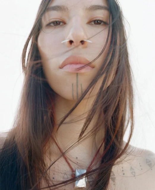 Close-up portrait of a young adult woman with long dark hair, nose piercing, and facial tattoo against a bright, out-of-focus background.