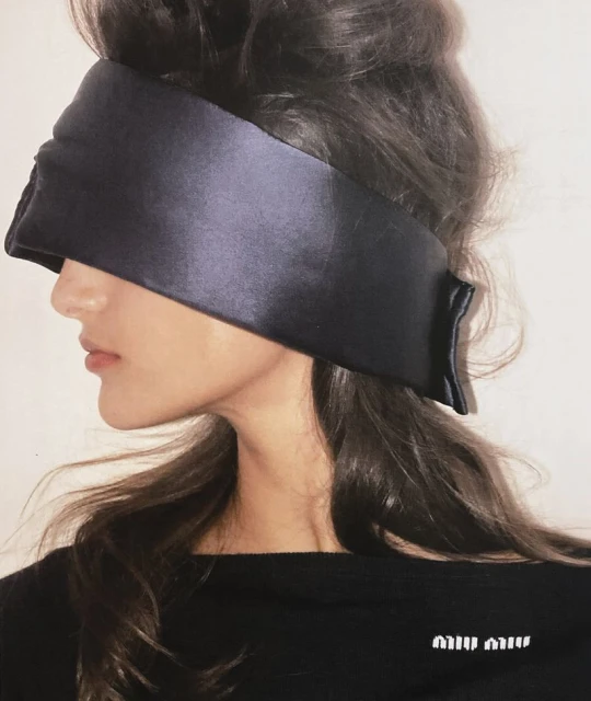 Woman with sleek, dark hair and a black satin blindfold poses in a profile shot wearing a black top with the Miu Miu logo.