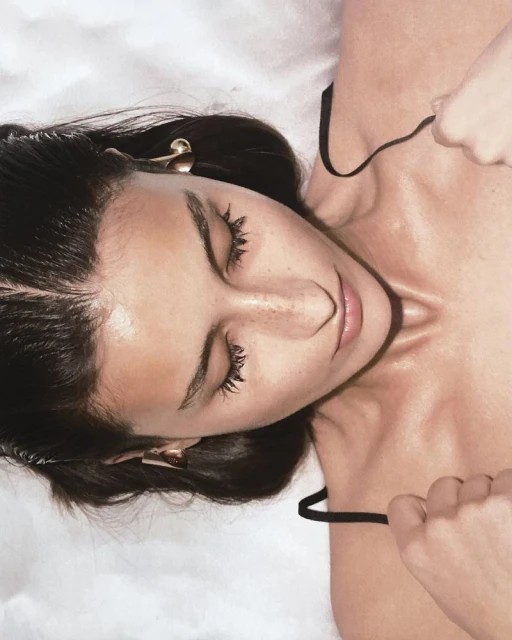 Close-up portrait of a woman with dark hair lying on a white surface, showcasing beauty makeup and subtle skin tones in soft lighting.
