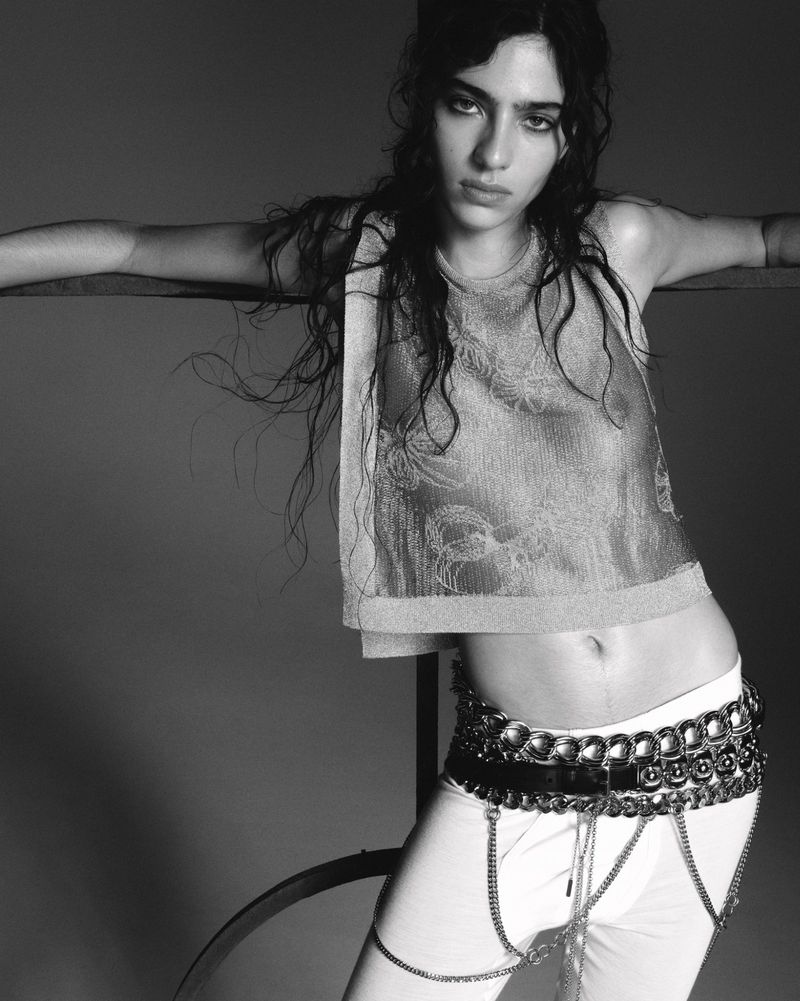 Black and white portrait of a young woman with wavy hair wearing a crop top, pants, and statement belt, striking a casual pose.