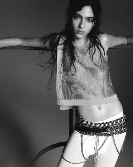 Black and white portrait of a young woman with wavy hair wearing a crop top, pants, and statement belt, striking a casual pose.