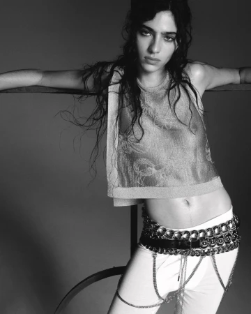 Black and white portrait of a young woman with wavy hair wearing a crop top, pants, and statement belt, striking a casual pose.