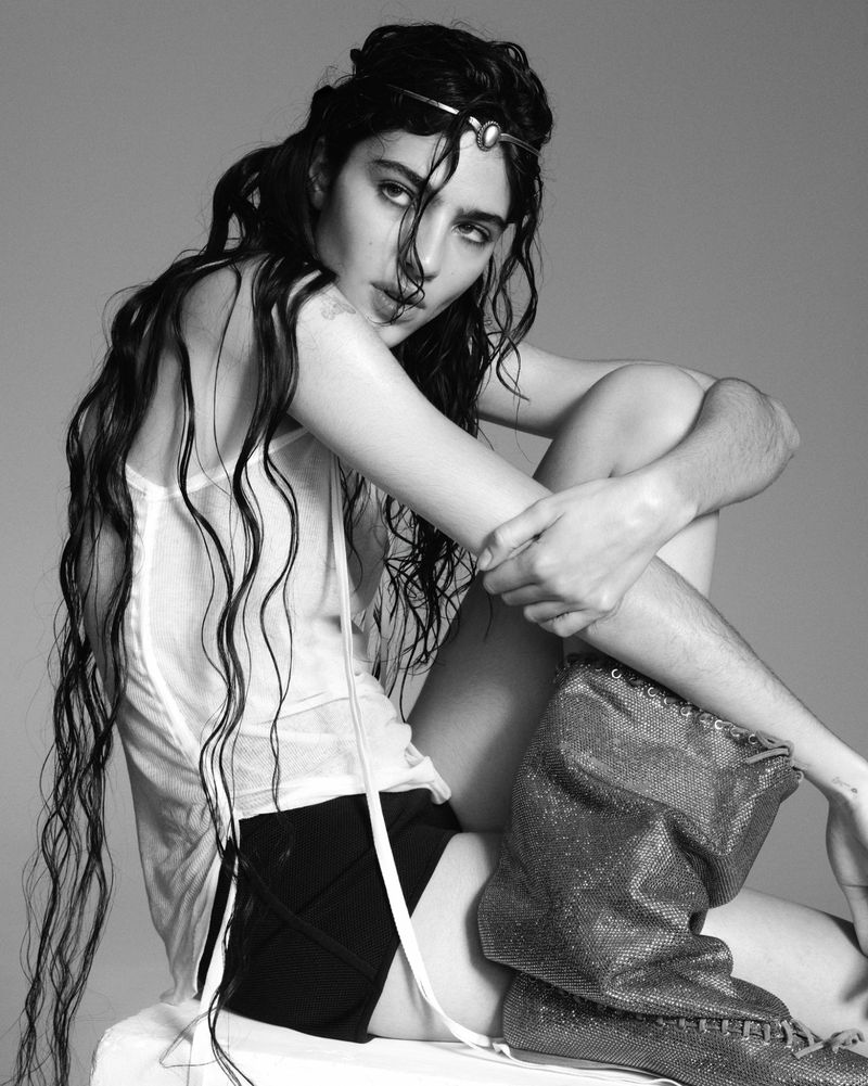 A woman with long dark hair and silver headpiece poses in a studio wearing a white top and black shorts with silver metal boots in black and white.