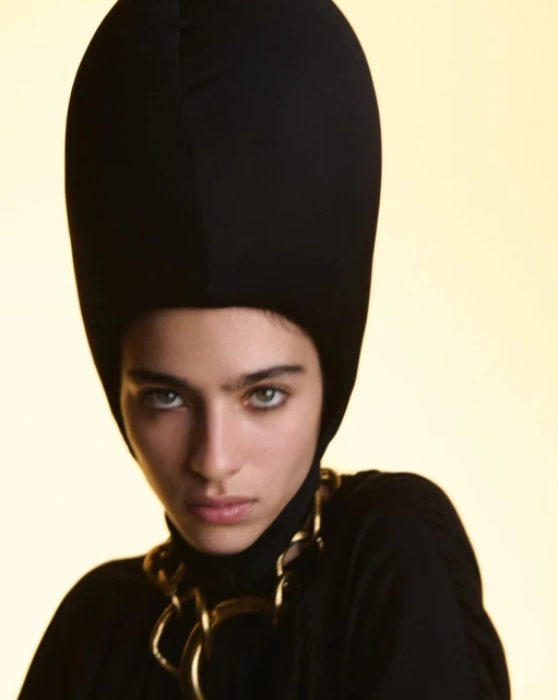 Close-up portrait of a woman wearing a black turtleneck top, a black balaclava-style hat, and a large gold chain necklace against a yellow background.