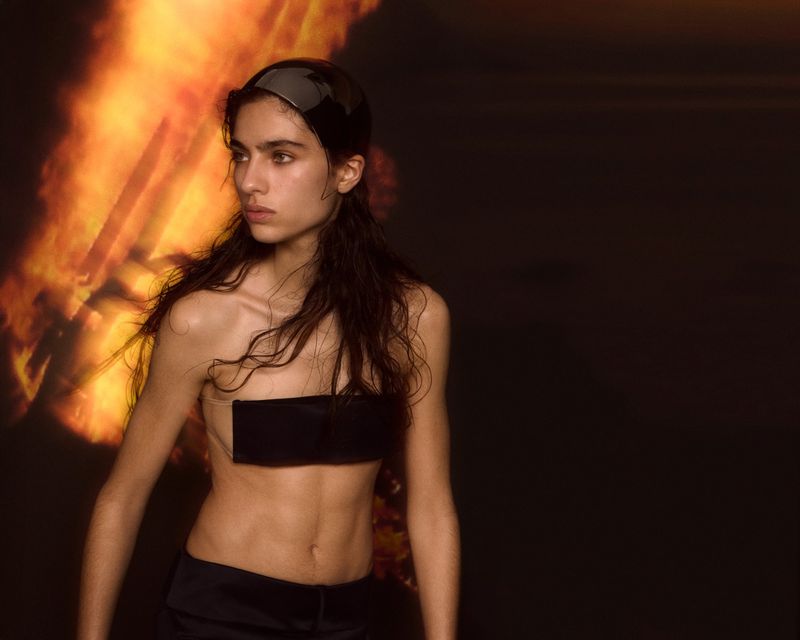 Portrait of a model with wet hair, wearing a black bandeau top and head covering, posing in front of a dark backdrop with red and orange light.