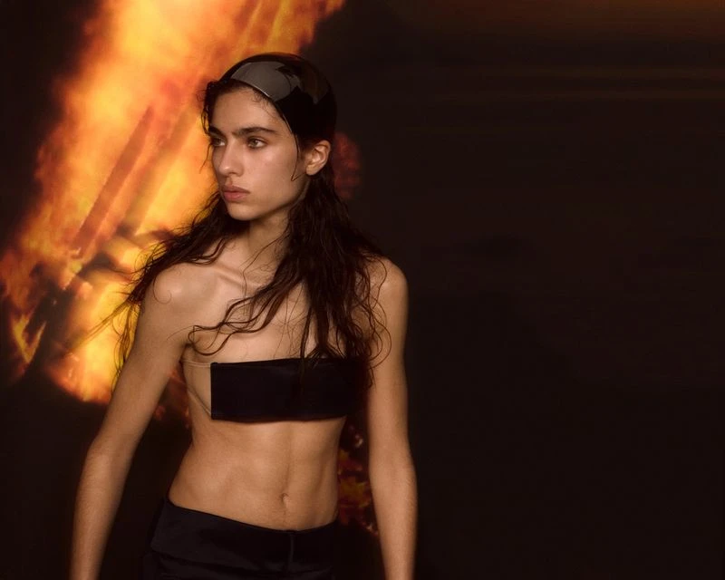 Portrait of a model with wet hair, wearing a black bandeau top and head covering, posing in front of a dark backdrop with red and orange light.