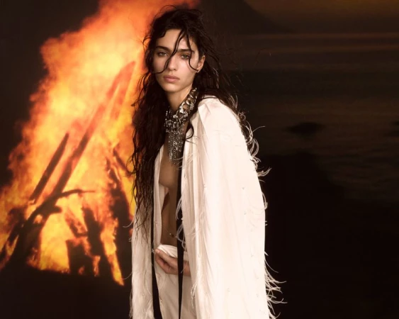 Adult female in a white fringed shawl poses with a silver necklace against a backdrop featuring a nighttime ocean and burning sticks.