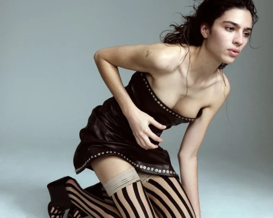 Brunette woman kneeling in a black studded dress and striped stockings against a grey backdrop in a studio fashion shot.